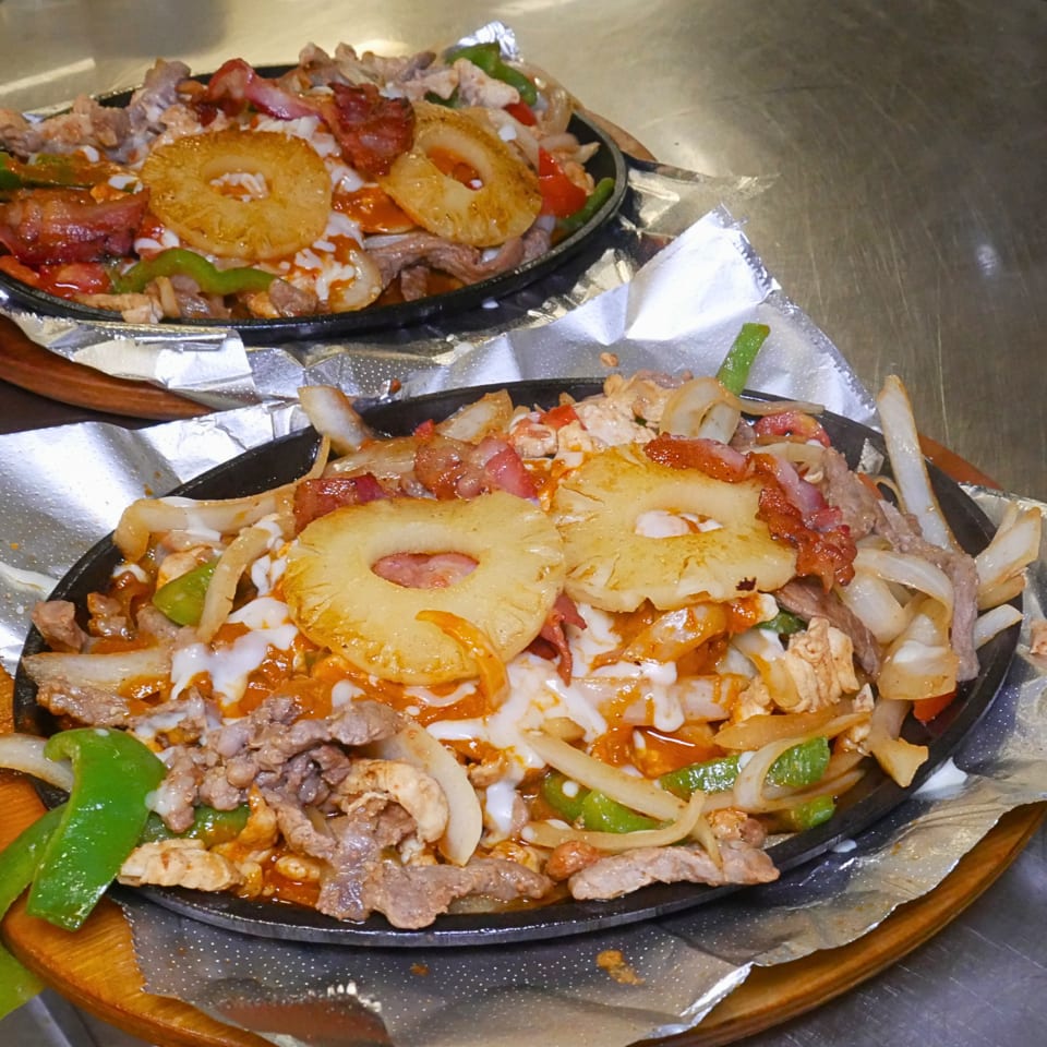 Sizzling mixed-grill platters topped with pineapple, cheese, peppers, onions, and red sauce on foil-lined wooden boards.