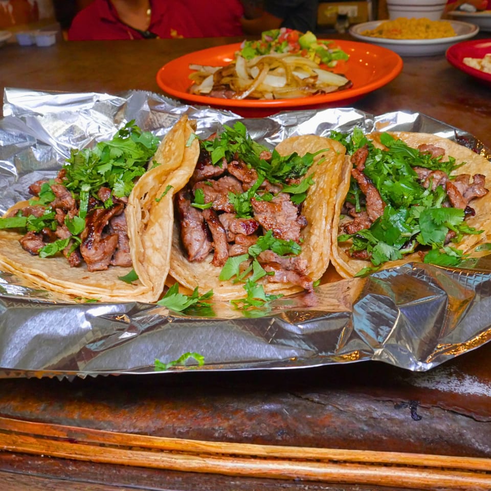 Grilled beef tacos topped with fresh cilantro on foil, with nachos and salsa visible on a restaurant table.