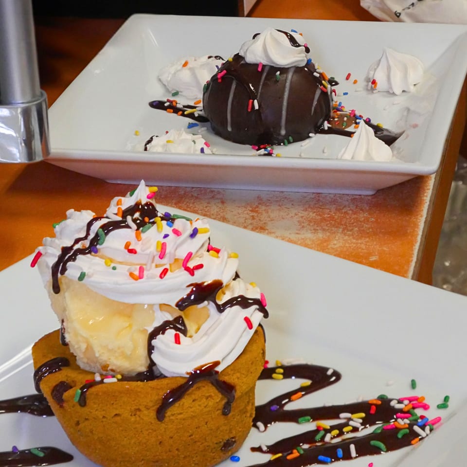 Dessert plate topped with whipped cream, chocolate sauce, colorful sprinkles, and a chocolate-covered cake reflected in a mirror.