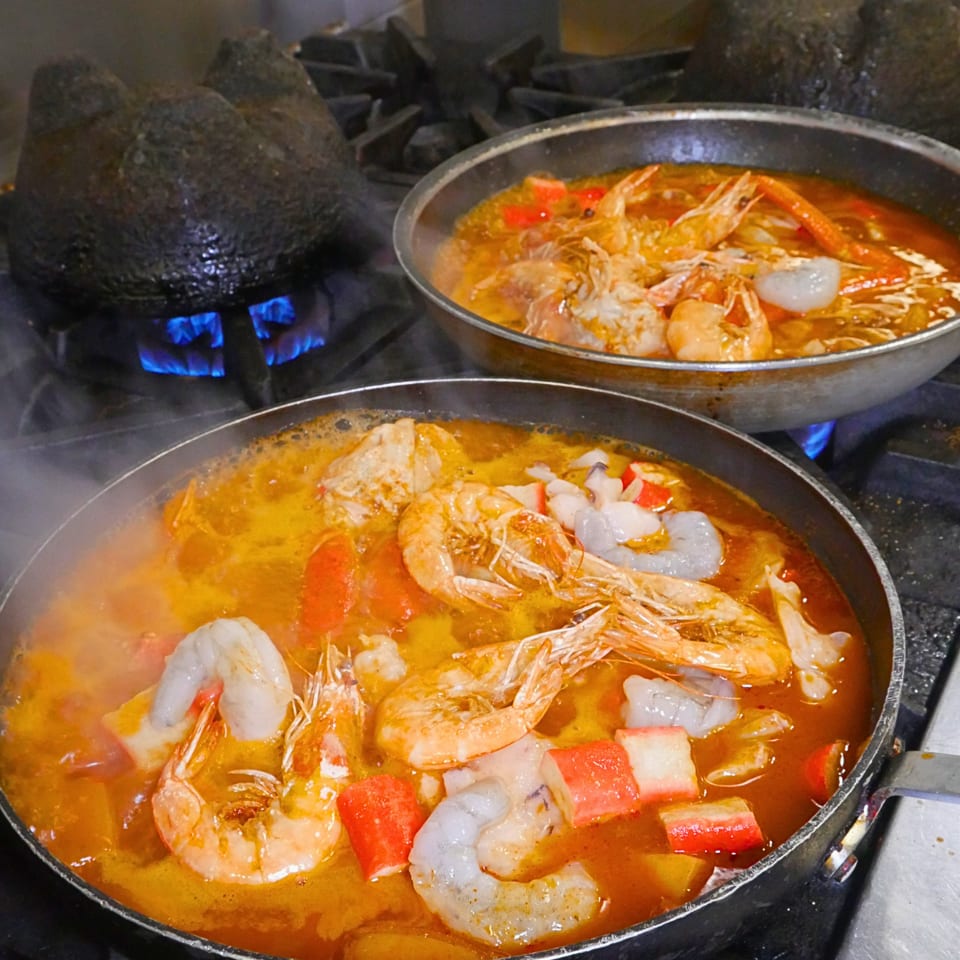 Two pans of spicy seafood soup with shrimp, squid, crab sticks, and vegetables cooking over blue gas flames.
