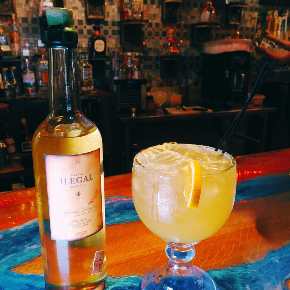 Mezcal bottle beside a salted-rim citrus cocktail with ice and a lime wedge on a colorful bar.