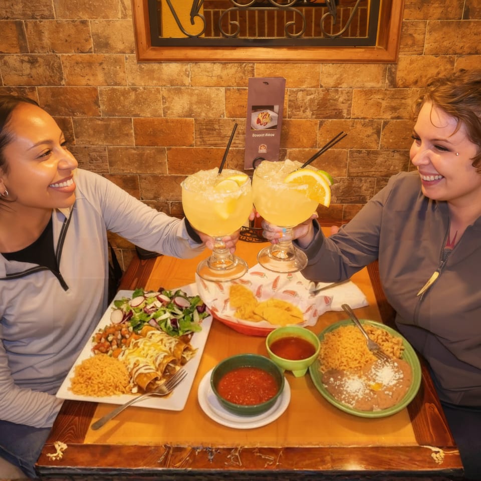 Two women toast with margaritas over a table of Mexican food in a warmly lit restaurant.