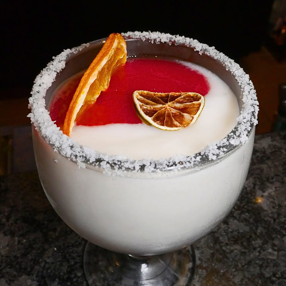 Red cocktail with a salt-rimmed glass, garnished with an orange peel and dried citrus slice.