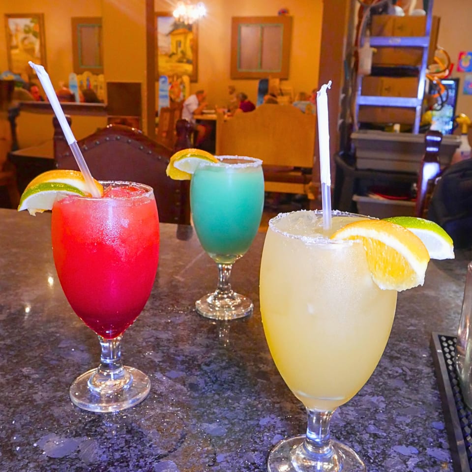 Three colorful margaritas with citrus garnishes and straws sit on a restaurant table.
