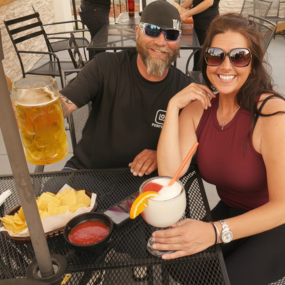 Smiling couple in sunglasses share drinks, tortilla chips, and salsa at an outdoor restaurant patio table.