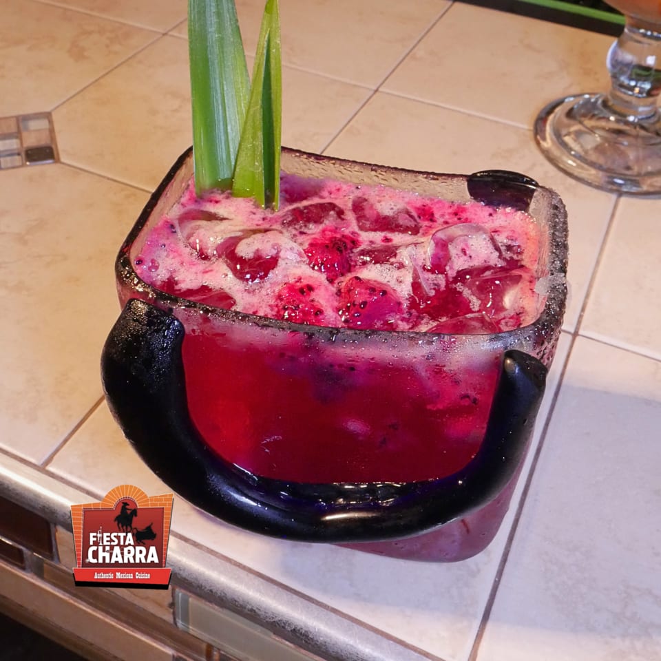 Vibrant pink fruit cocktail with ice, green leaves, and a black straw in a decorative glass at Fiesta Charra.