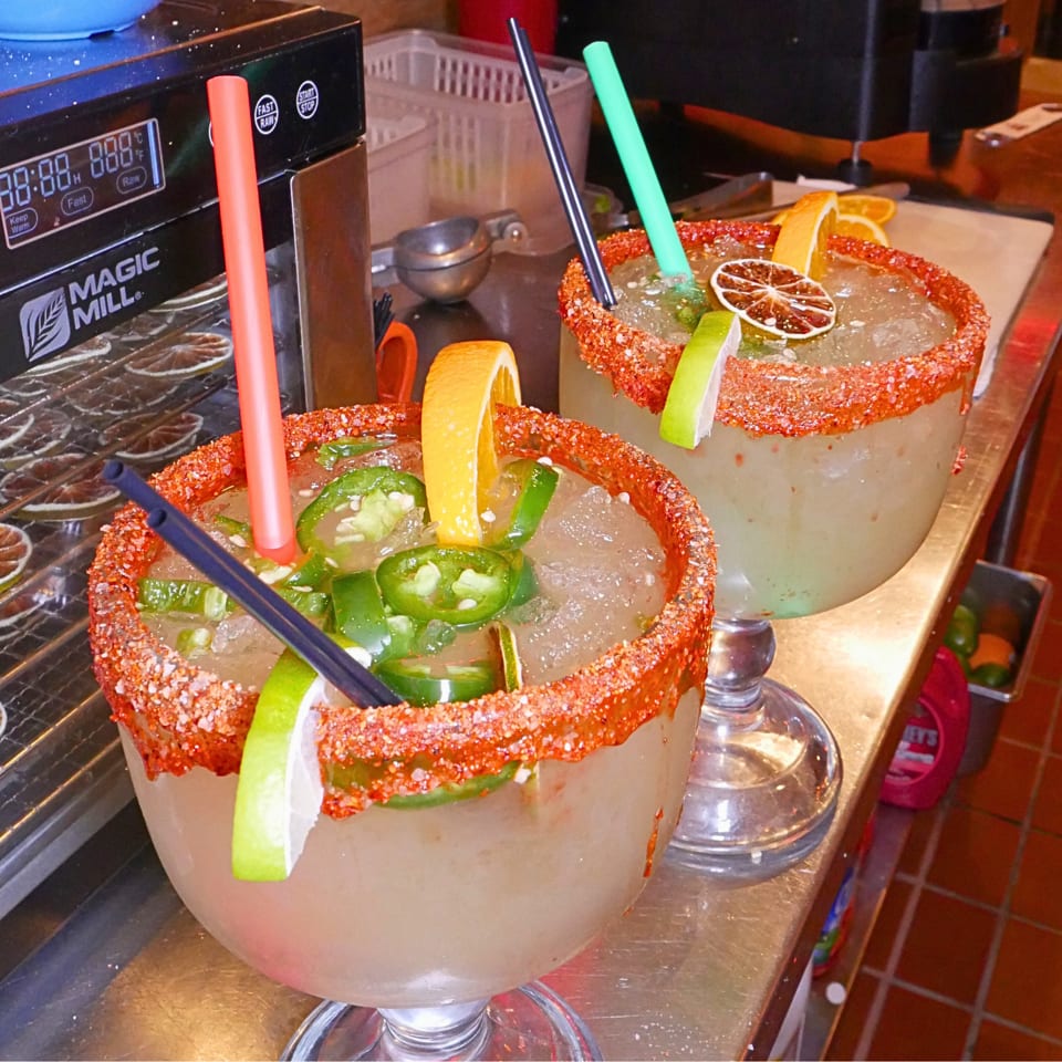 Two spicy cocktails with red chili-salt rims, jalapeño slices, citrus garnishes, and colorful straws on a restaurant bar.