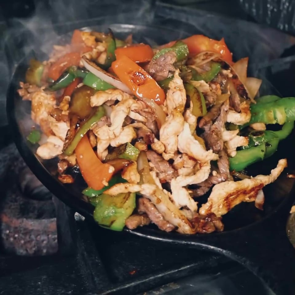 Sizzling stir-fry with grilled chicken, beef, carrots, green peppers, zucchini, and onions in a black pan.