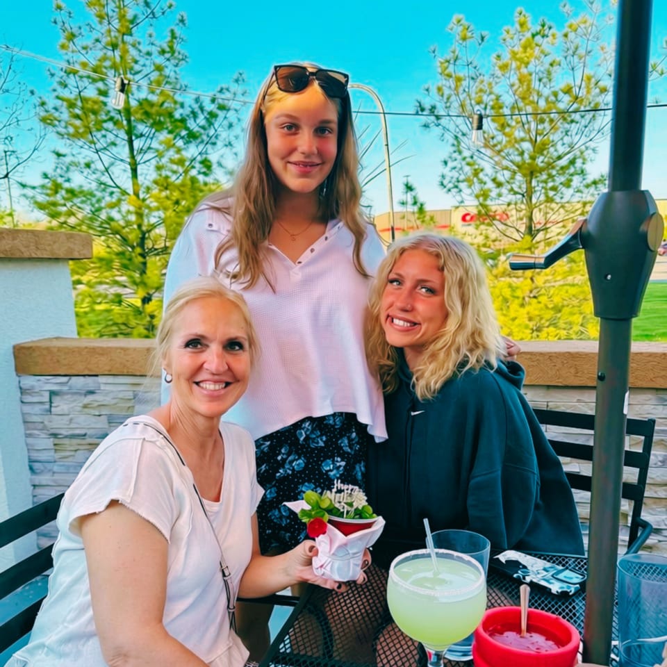 Three smiling women pose at an outdoor restaurant table with drinks and a flower-topped dessert.