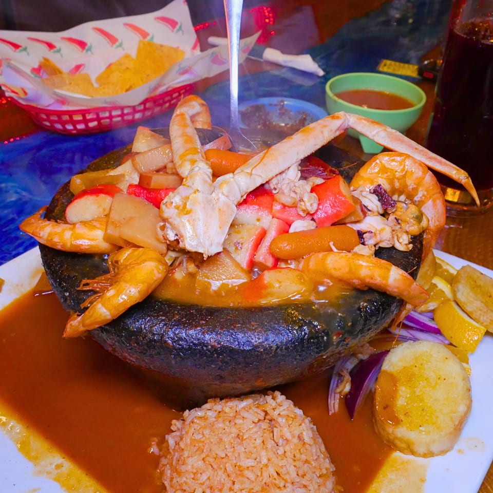 Mixed seafood platter with crab legs, shrimp, mussels, vegetables, rice, and dipping sauce on a sizzling black stone plate