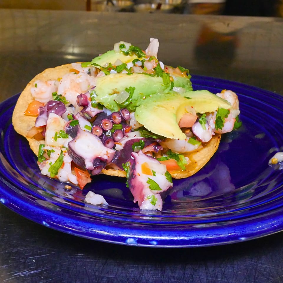 Octopus ceviche tostada topped with avocado, red onion, tomato, and cilantro on a blue plate.
