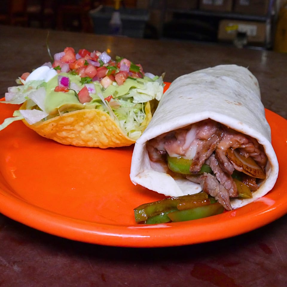 Orange plate holding a steak-filled tortilla wrap with peppers and a taco topped with lettuce, avocado, tomato, and onion.