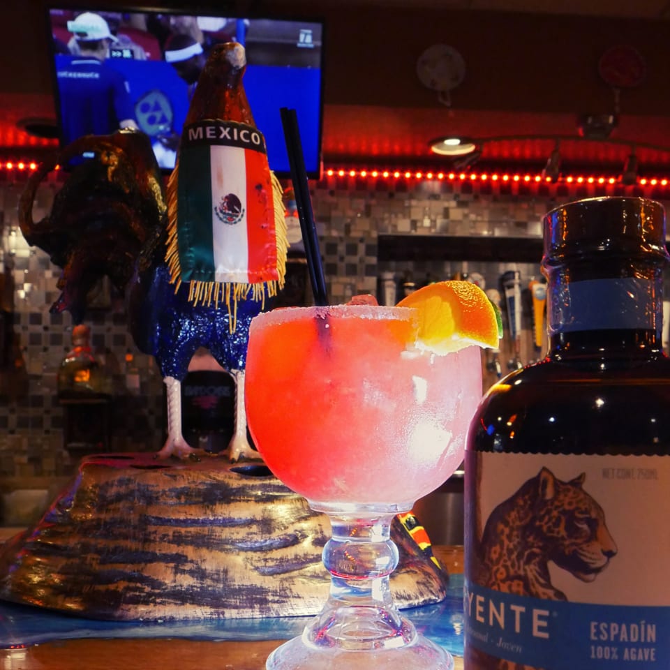 Pink margarita with a salted rim and lime wedge beside Mexican tequila bottles in a warmly lit restaurant bar.
