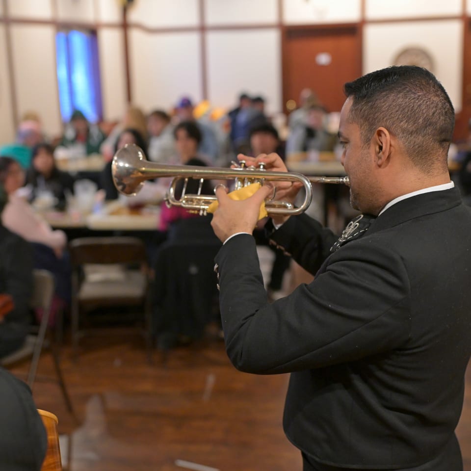Musician in a black suit plays trumpet to diners in a warmly lit restaurant.